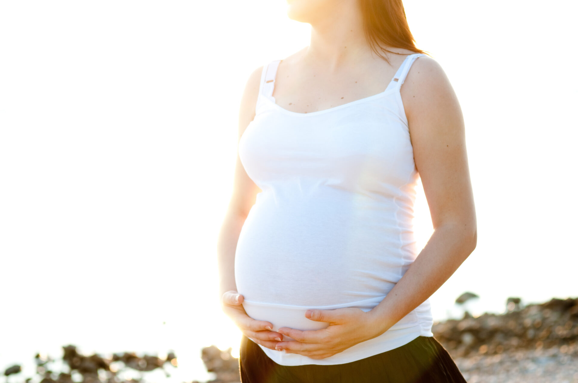 Close up of a pregnant woman in a white tank top with the sun shining behind her.